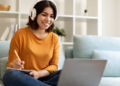 Jovem estudando online com fones de ouvido e anotando no caderno.
