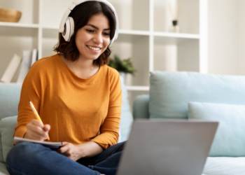 Jovem estudando online com fones de ouvido e anotando no caderno.
