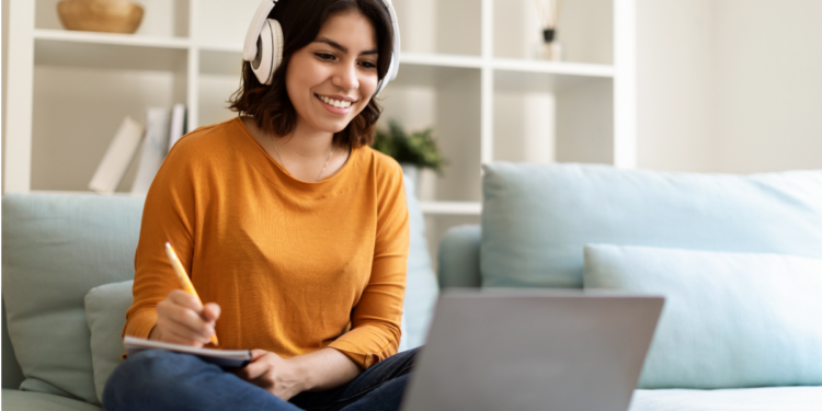 Jovem estudando online com fones de ouvido e anotando no caderno.