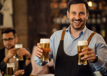 Homem sorridente segurando copos de cerveja em bar.