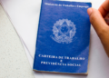 Carteira de Trabalho azul sobre mesa ao lado de mão segurando caneta.
