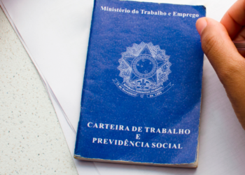 Carteira de Trabalho azul sobre mesa ao lado de mão segurando caneta.