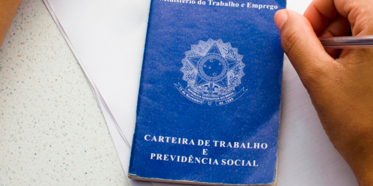 Carteira de Trabalho azul sobre mesa ao lado de mão segurando caneta.