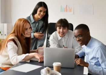 Um grupo de quatro jovens profissionais reunidos ao redor de um notebook, sorrindo enquanto acompanham juntos o conteúdo exibido na tela.