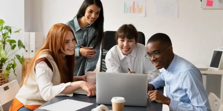 Um grupo de quatro jovens profissionais reunidos ao redor de um notebook, sorrindo enquanto acompanham juntos o conteúdo exibido na tela.