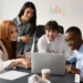 Um grupo de quatro jovens profissionais reunidos ao redor de um notebook, sorrindo enquanto acompanham juntos o conteúdo exibido na tela.