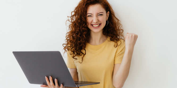 Jovem comemora vitória com notebook em fundo claro.