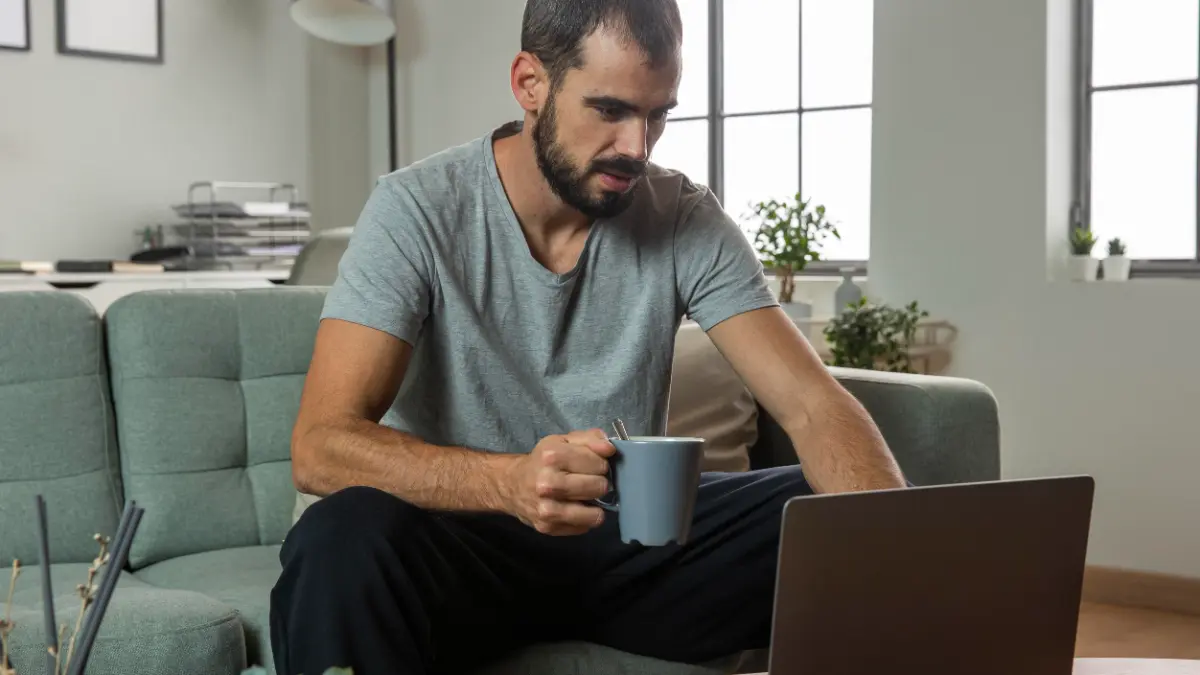Homem em ambiente doméstico mexendo no notebook e segurando caneca.