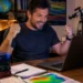 Homem comemora sorrindo diante do notebook em seu ambiente de estudos, cercado por livros, mapas, globo terrestre e instrumentos científicos.