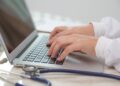 Mãos de médico digitando em notebook com estetoscópio azul sobre mesa clara ao lado, simbolizando o acesso aos locais de prova do Enamed 2026 e da primeira etapa do Revalida 2026/2.