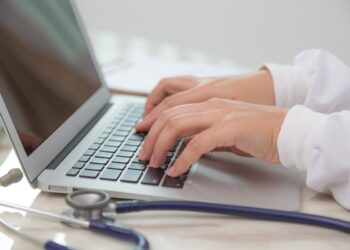 Mãos de médico digitando em notebook com estetoscópio azul sobre mesa clara ao lado, simbolizando o acesso aos locais de prova do Enamed 2026 e da primeira etapa do Revalida 2026/2.