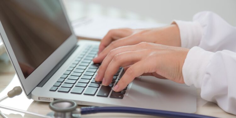 Mãos de médico digitando em notebook com estetoscópio azul sobre mesa clara ao lado, simbolizando o acesso aos locais de prova do Enamed 2026 e da primeira etapa do Revalida 2026/2.