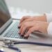 Mãos de médico digitando em notebook com estetoscópio azul sobre mesa clara ao lado, simbolizando o acesso aos locais de prova do Enamed 2026 e da primeira etapa do Revalida 2026/2.