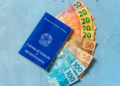 Carteira de Trabalho azul encostada em notas de dinheiro real brasileiro e fundo azul.