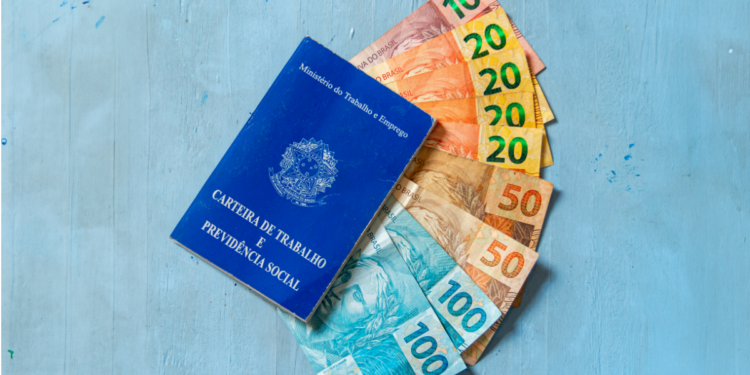 Carteira de Trabalho azul encostada em notas de dinheiro real brasileiro e fundo azul.