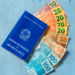 Carteira de Trabalho azul encostada em notas de dinheiro real brasileiro e fundo azul.