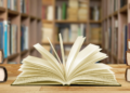 Livro aberto sobre mesa em biblioteca com estantes ao fundo.