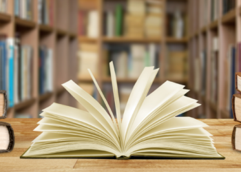 Livro aberto sobre mesa em biblioteca com estantes ao fundo.