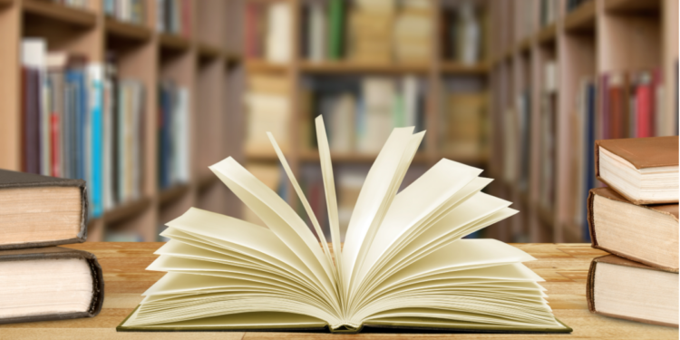 Livro aberto sobre mesa em biblioteca com estantes ao fundo.