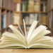 Livro aberto sobre mesa em biblioteca com estantes ao fundo.