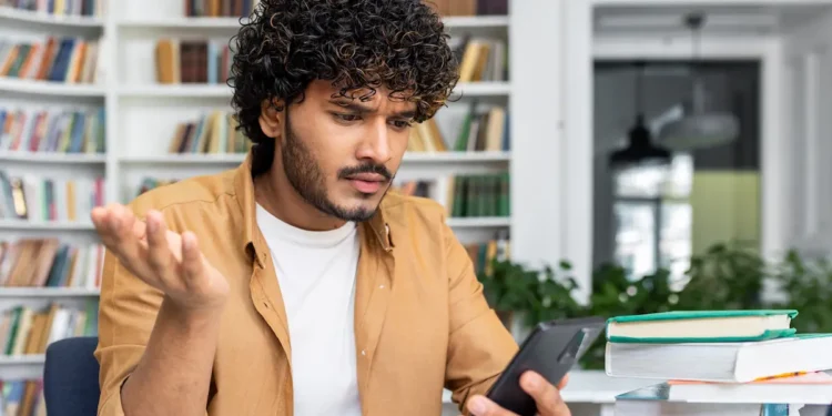 Estudante consulta o celular com expressão de dúvida sobre pagamento do Pé-de-Meia de agosto.