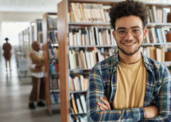 Jovem sorridente de óculos em biblioteca universitária moderna.