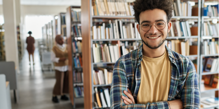 Jovem sorridente de óculos em biblioteca universitária moderna.