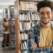 Jovem sorridente de óculos em biblioteca universitária moderna.