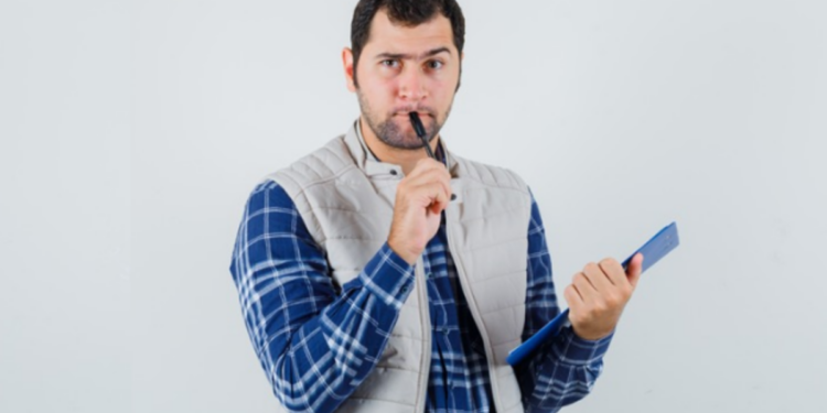 Homem segurando prancheta e caneta com expressão pensativa sobre fundo branco.