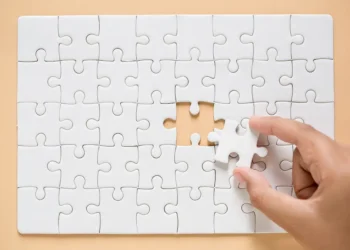 Mão encaixando peça em quebra-cabeça branco sobre fundo bege.