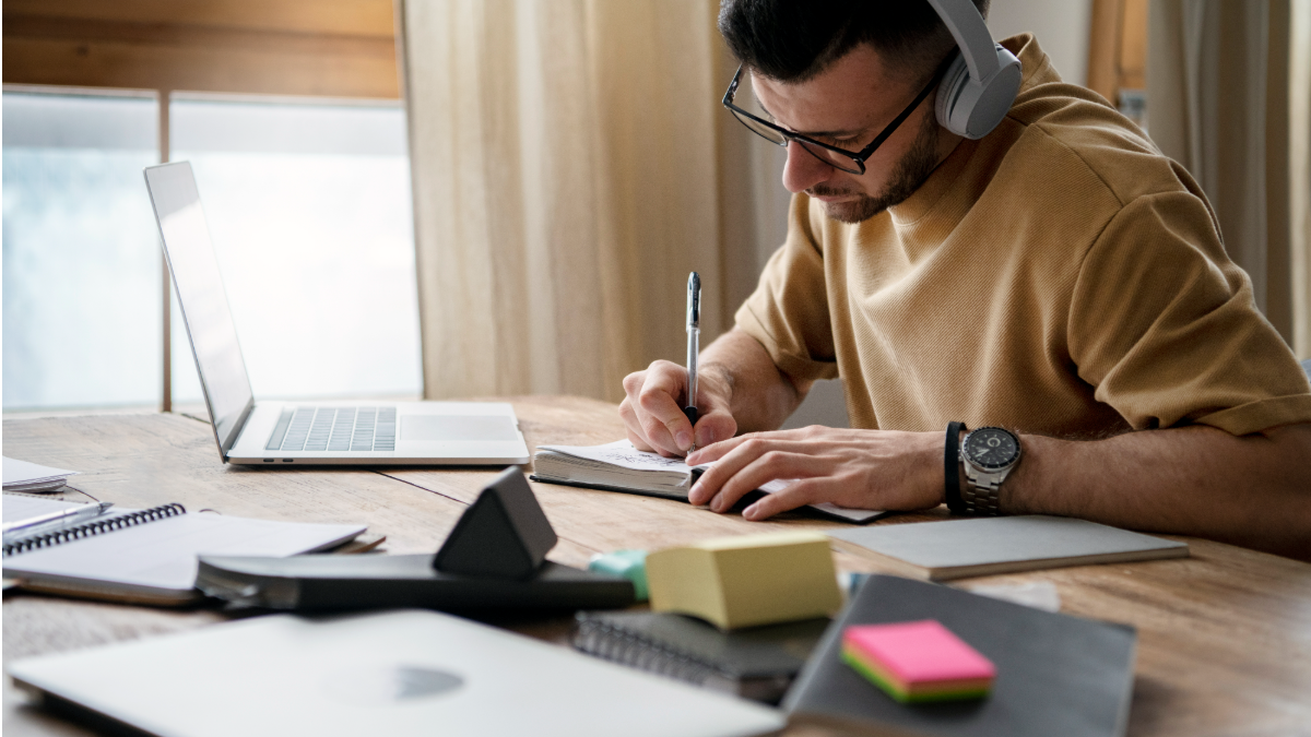 Jovem estudando com fones de ouvido e laptop em mesa de madeira.