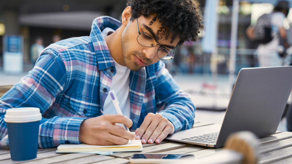 Jovem estudando ao ar livre com notebook e caderno em ambiente urbano.