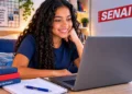 Estudante sorridente acompanha uma aula pelo notebook em um quarto aconchegante e organizado, com o logo do SENAI em destaque ao fundo.