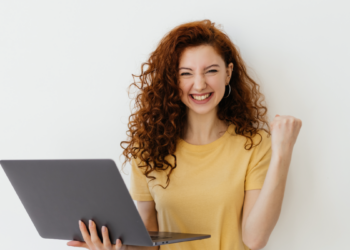 Jovem comemora vitória com notebook sorrindo e punho cerrado.
