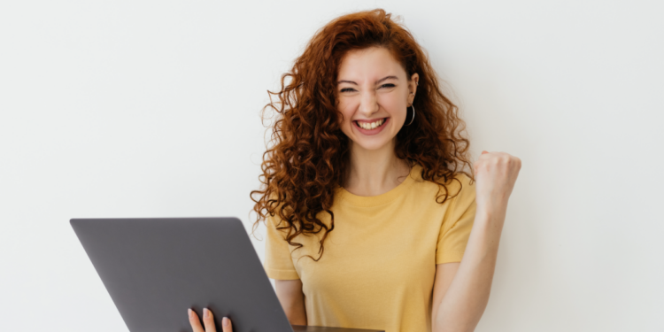 Jovem comemora vitória com notebook sorrindo e punho cerrado.