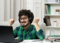 Homem sorridente com fones levanta os punhos de frente ao notebook em ambiente doméstico.