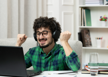 Homem sorridente com fones levanta os punhos de frente ao notebook em ambiente doméstico.