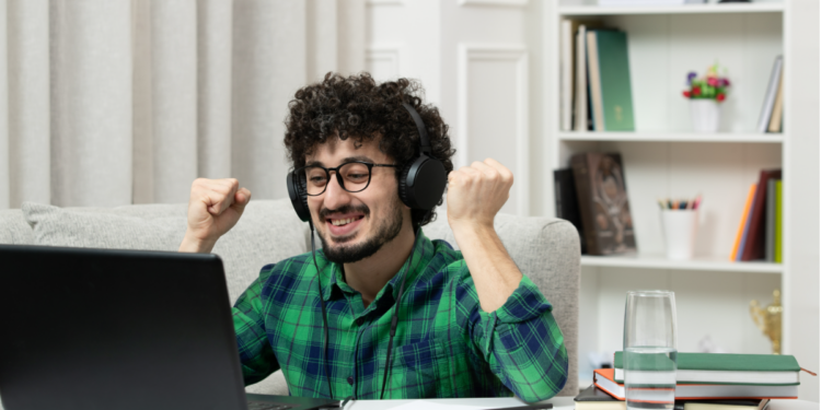 Homem sorridente com fones levanta os punhos de frente ao notebook em ambiente doméstico.