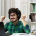 Homem sorridente com fones levanta os punhos de frente ao notebook em ambiente doméstico.