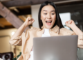 Mulher jovem com expressão de euforia levanta os braços em frente a notebook prata em sala iluminada, simbolizando as oportunidades de emprego no Rio de Janeiro.