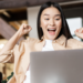 Mulher jovem com expressão de euforia levanta os braços em frente a notebook prata em sala iluminada, simbolizando as oportunidades de emprego no Rio de Janeiro.