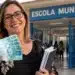 Professora segurando cédulas de dinheiro em frente a uma escola municipal, representando o anúncio do abono de R$ 3.500 para profissionais da educação.