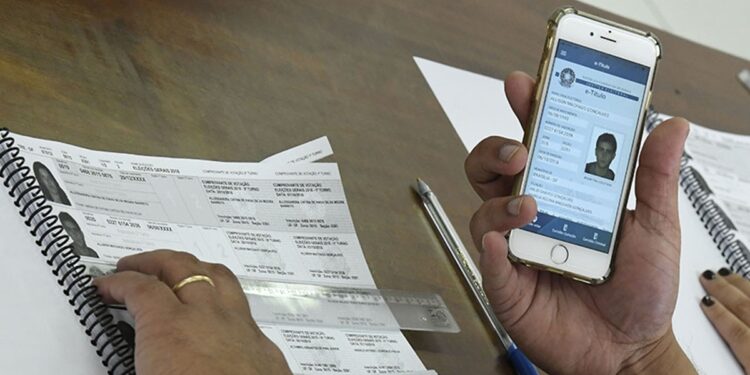 Imagem ilustrativa: mesário confere o caderno de votação enquanto um eleitor apresenta o e-Título na tela do celular