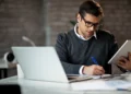 Pessoa atuando como analista em ambiente de trabalho, utilizando notebook e tablet para análise de informações e planejamento profissional.