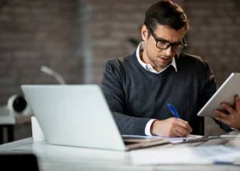 Pessoa atuando como analista em ambiente de trabalho, utilizando notebook e tablet para análise de informações e planejamento profissional.