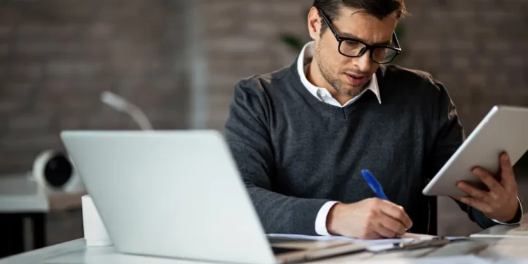 Pessoa atuando como analista em ambiente de trabalho, utilizando notebook e tablet para análise de informações e planejamento profissional.