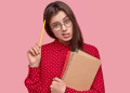 Imagem ilustrativa de jovem de óculos redondos com expressão de dúvida, coçando a cabeça com um lápis e segurando cadernos, em referência ao aumentativo de festa