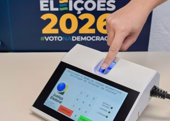 Imagem ilustrativa de dedo posicionado no leitor biométrico do terminal do mesário, com painel das Eleições 2026 ao fundo, em referência à biometria nas Eleições 2026