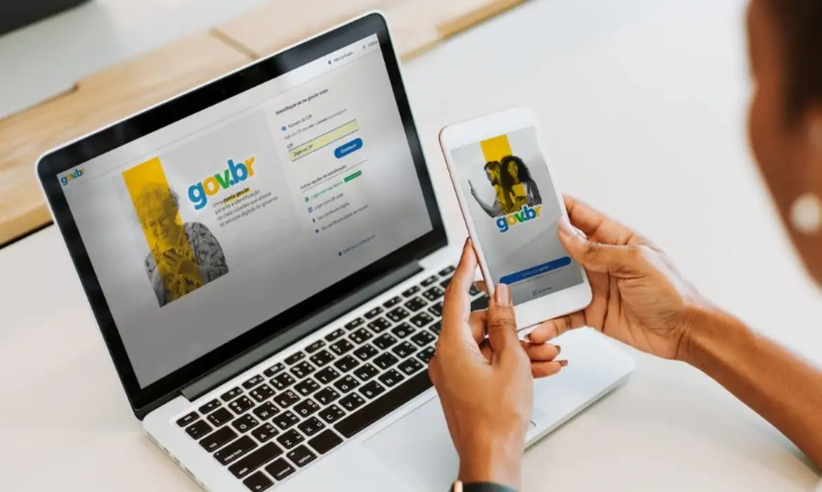 Imagem ilustrativa de mãos segurando celular com o aplicativo gov.br aberto, diante de um notebook com a tela de login da plataforma, em referência à Carteira de Identidade Nacional