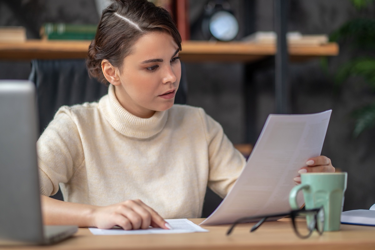 Jovem mulher lê atentamente apostila ao lado de notebook e caneca em mesa de estudos, simbolizando a leitura da cartilha do Enem.
